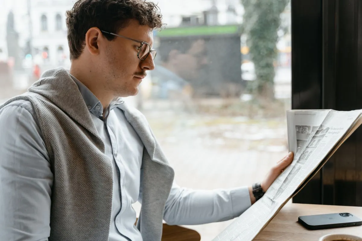 Un homme portant des lunettes lit un journal près d’une fenêtre